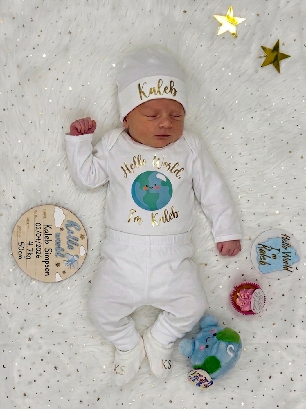 Newborn baby in a white outfit with 'Hello World' and 'Kaleb' text, lying on a textured surface.